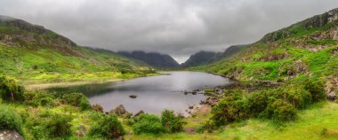 Dunloe Geçidi 'nin yemyeşil tepeleri arasında zarif bir şekilde yerleşmiş, huzurlu bir göl manzarası hayal edin. Yumuşak bulutlarla kaplanmış, huzurlu bir atmosfer yaratıyor, Kerry, İrlanda