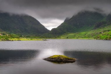 Huzurlu ve huzurlu bir dağ gölü, Dunloe Geçidi 'nin canlı yeşilliği içinde, dramatik, pofuduk bulutların altında, doğanın huzur veren güzelliğini güzel bir şekilde yansıtıyor, Kerry, İrlanda
