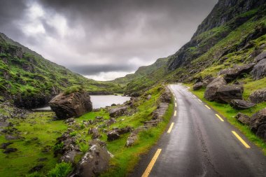 Büyüleyici bir yol, yemyeşil tepeler ve engebeli Dunloe kayalık arazisinde dramatik, karamsar gökyüzünün altında manzara fotoğrafçılığı için mükemmel bir zemin oluşturur, Kerry, İrlanda