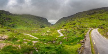 Bulutlu bir gökyüzünün altındaki görkemli dağlık yamaçlar tarafından güzelce çerçevelenmiş Dunloe Vadisi 'nin yemyeşil bir geçidinde zarifçe ilerleyen dolambaçlı bir yolun huzurlu ve huzurlu manzarası.
