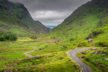 Dolambaçlı bir yol zarif bir şekilde Dunloe Vadisi 'nin verimli ve canlı bir geçidini kesiyor. Etkileyici bir gökyüzünün altındaki dramatik, yüksek dağlarla çevrili, büyüleyici bir manzara yaratıyor, Kerry, İrlanda