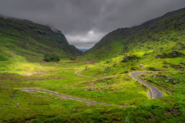 Etrafı görkemli tepeler ve karamsar bulutlarla çevrili, kıvrımlı yeşil Dunloe vadisinde zarif bir şekilde örülmüş bir yol, gerçekten sakin ve resimsel bir manzara yaratıyor, Kerry, İrlanda