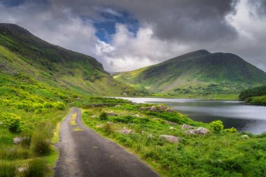 Manzaralı bir yol, yemyeşil bir göle doğru esiyor. Kara Vadi 'deki görkemli dağlar tarafından çerçevelenmiş, dramatik bir gökyüzünün altında büyüleyici bir doğal manzara yaratıyor, Kerry, İrlanda.