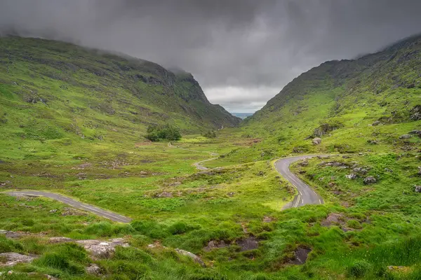 Huzurlu ve pitoresk yeşil bir vadi Dunloe Geçidi 'nin görkemli tepeleri ve bulutlu gökyüzü tarafından kucaklanan dolambaçlı bir yol sergiliyor, doğanın huzurunu kusursuz bir şekilde yansıtan Kerry, İrlanda