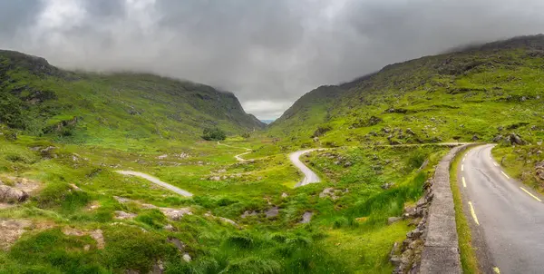 Bulutlu bir gökyüzünün altındaki görkemli dağlık yamaçlar tarafından güzelce çerçevelenmiş Dunloe Vadisi 'nin yemyeşil bir geçidinde zarifçe ilerleyen dolambaçlı bir yolun huzurlu ve huzurlu manzarası.