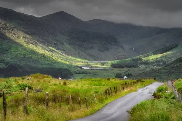 Yuvarlanan dağların nefes kesici manzarasını keşfedin ve keşfedin ve huzur dolu bir Kara Vadi, bulutların arasından bakan sıcak güneş ışığıyla güzelce aydınlatılmış, Kerry, İrlanda