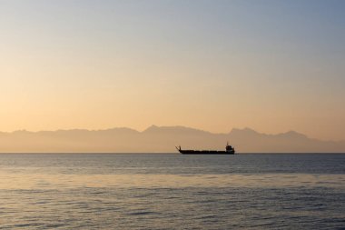 Bir kargo mavnası şafak vakti denizde, sisli, kargo ve yolcu taşımacılığında dağların arka planına karşı yüzer.. 
