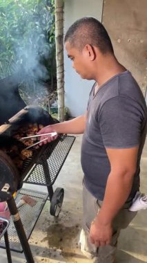 Bir adam döner ve ızgarada maşayla eti değiştirir ateşte ızgara yapar, barbekü pişirir. 