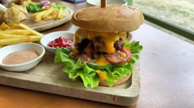 Salata ve sebzeli büyük, sulu bir burger, patates kızartması ve sosla servis edilir. Pencere kenarındaki bir kafede, ahşap bir tepside.