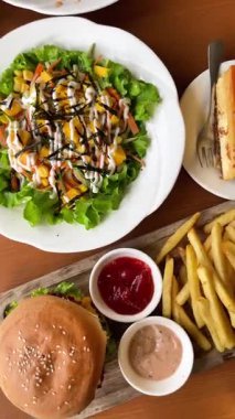 Salata ve sebzeli büyük, sulu bir burger, patates kızartması ve sosla servis edilir. Masadaki bir kafede, ahşap tepside ve peynirli kek ile..