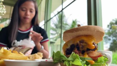 Genç bir kadın, arka planda büyük sulu hamburger ve soslar eşliğinde restoranda milkshake içiyor.