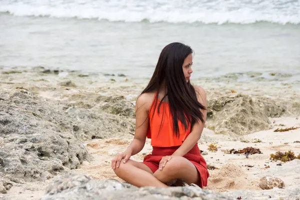 Genç, güzel Asyalı, dalgalı, uzun, esmer saçlı, mayo giymiş bir kadın. Kayalık sahilde deniz kenarında oturuyor..