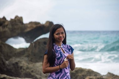 Arkasında dalgalarla tropik sahilde poz veren bir kadın..