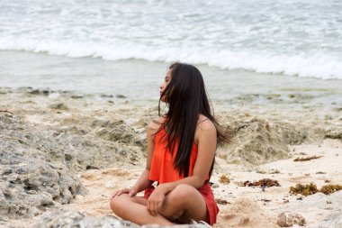 Genç, güzel Asyalı, dalgalı, uzun, esmer saçlı, mayo giymiş bir kadın. Kayalık sahilde deniz kenarında oturuyor..