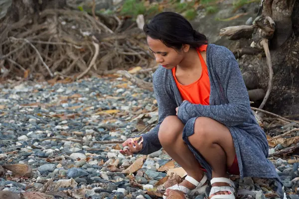 Genç Asyalı kadın tropik kökler yakınlarındaki çakıl taşı plajında deniz kabuğu topluyor..