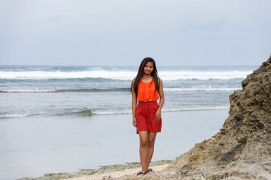 Okyanusun kenarında duran genç bir kadın. Arkasında kayalar, deniz hiddetli..