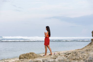 Genç bir kadın, elleri duada katlanmış olarak denizin önünde duruyor ve fırtınalı denize bakıyor..