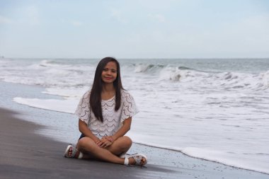 Genç kadın dalgaların yanında kumsalda oturuyor..