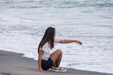 Genç Asyalı kadın gülümsüyor, kumlu sahilde deniz kenarında oturuyor..