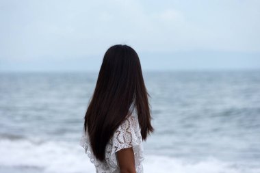 Genç bir kadın kıyıda tek başına duruyor ve deniz dalgalarına bakıyor..
