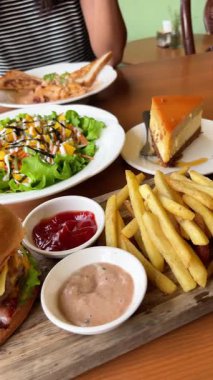 Salata ve sebzeli büyük, sulu bir burger, patates kızartması ve sosla servis edilir. Masadaki bir kafede, ahşap tepside ve peynirli kek ile..