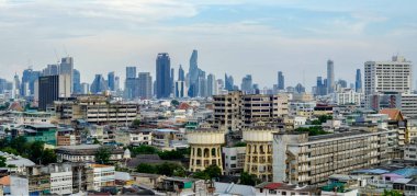 Bangkok, Tayland 'ın geniş açılı panoramik manzarası, arka planda çürüyen orta yüzyıl binaları ile modern yüksek binalar arasında çarpıcı bir kontrastla karşılaşıyor. Görüntü, Tayland başkentinin gelişmekte olan kentsel manzarasını net ayrıntılarla vurguluyor