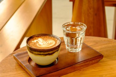 Bir fincan kahvenin ve güzel bir latte sanatının, her ikisi de ahşap bir tepside duran temiz bir bardak suyun yanında sıcacık bir şekilde aydınlatılmış bir hayat sahnesi. Bu resim, kahve hazırlama ve tazeleme sanatını vurgulayan tipik bir kafe sahnesini yakalar.