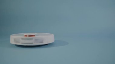 Beyaz otomatik elektrikli süpürge, açık mavi stüdyo arka planında izole edilmiş zemini temizler. Robot elektrikli süpürge masayı temizliyor, akıllı algılayıcılar yüksek kalite 4K görüntü