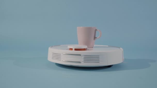 コーヒーカップが独立した青の背景を持つスタジオで回転する自律型掃除機 高品質4K映像 — ストック動画