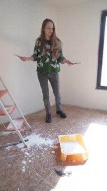Neşeli bir kadın resim yaparken, fırça ve silindirle boya dökerken darmadağın olur. DIY, ev yenileme ve eğlenceli yaratıcı projeler için mükemmel stok görüntüleri..