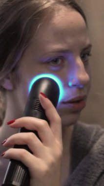 Bu dikey videoyla son moda güzellik akımları. Bir kadın aynanın önüne LED yüz aygıtıyla cilt bakımı uyguluyor. Modern bir banyoda geçiyor. Güzellik reklamları için mükemmel.