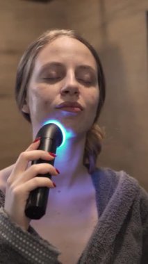 Modern bir banyoda LED yüz aygıtı kullanan bir kadının dikey videosuyla kendine bakım anı. Güzellik dersleri, yaşam tarzı terfileri ve gelişmiş cilt bakımı için ideal.