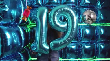  19 numaralı mavi folyo balonunu elinde tutan genç bir kadın, canlı bir disko arka planına karşı özel bir doğum gününü veya yıldönümünü kutluyor..