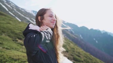 Genç bir kadın, rüzgarın saçlarının arasında yavaşça hareket ederken gülümsüyor. Arka planda görkemli bir dağ manzarası var. Bu yavaş çekim bir neşe, özgürlük ve doğal güzelliği yakalar.