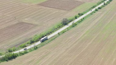 Bulgaristan 'ın ilkbaharında yemyeşil tarım sahalarının etrafını sardığı dolambaçlı kırsal bir yoldan geçen ağır bir nakliye aracının sinematik hava aracı görüntüsü.