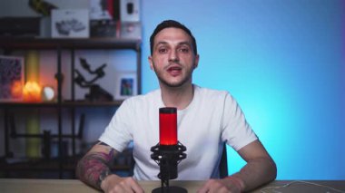 Dövmeli Avrupalı adam podcast stüdyosunda kameraya doğru konuşuyor. Yüksek kaliteli ışıklar, mikrofon ve profesyonel içerik oluşturucu kurulumu.