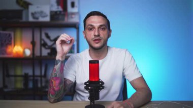 Dövmeleri olan kendine güvenen Avrupalı bir adam, yumuşak ışıklandırma ve mikrofon ayarlı profesyonel bir stüdyoda kamerayla konuşan bir podcast kaydediyor..