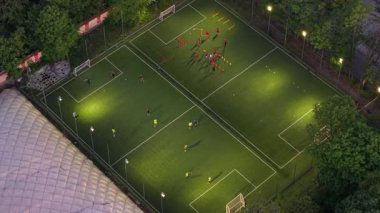 Yukarıdan aşağıya, yeşil ağaçlarla çevrili bir şehir parkında futbol maçını gösteren bir drone fotoğrafı. Sahnede oyuncuların ve aşağıda canlı yeşilliklerin olduğu sinematik kentsel doğa sahnesi.