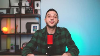 Genç erkek podcaster modern bir stüdyoda oturuyor, bir podcast ya da vlog bölümünü kaydederken güvenli bir şekilde mikrofona konuşuyor, profesyonel bir kurulumda kameraya bakıyor..