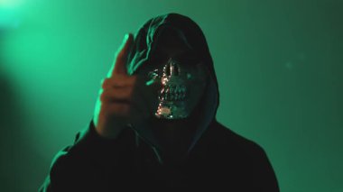 Kapüşonlu maskeli kişinin doğrudan kameraya bakıp karanlık odada çekildiği siber güvenlik tehdidi kavramı.