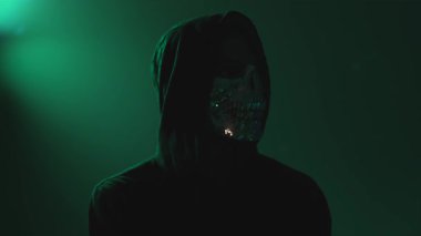 Kapüşonlu maskeli hacker karanlık odada otururken kameraya bakıyor, siber suç ve kimlik hırsızlığı konsepti, gizemli ve dramatik ruh hali