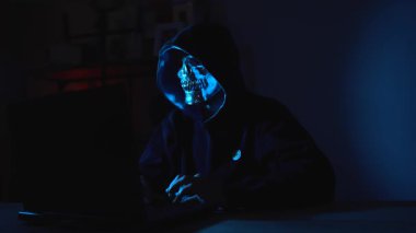 Maskeli şahıs karanlık odadaki dizüstü bilgisayarda yoğun bir şekilde yazıyor. Sayısal tehdit, bilişim güvenliği ve kimlik sahtekarlığı konuları için ideal hackleme, izleme, siber suç ve gizlilik ihlallerini temsil ediyor.