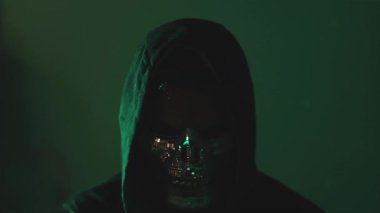 Karanlık odada kapüşonlu ve maskeli kamera izleyen şüpheli şahıs, hackleme ve internet dolandırıcılığının sembolü.