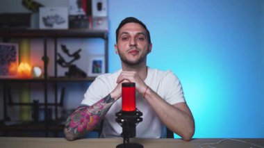 Dövmeli adam bir vlog ya da podcast oturumu sırasında doğrudan kameraya konuşur. Profesyonel donanımlı rahat stüdyo alanı.