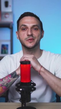 Profesyonel dövmeli bir erkek vlogger 'ın podcast kaydının dikey videosu. Avrupalı adam, YouTube 'da ışıklandırma ve mikrofon ile modern bir stüdyo ortamında kameraya konuşuyor..
