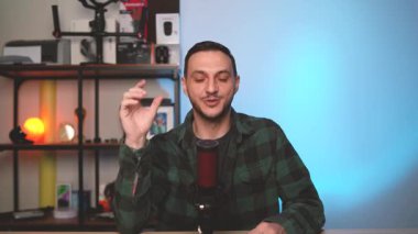 Kendine güvenen erkek vlogger modern bir stüdyoda podcast bölümü sunuyor, stüdyo mikrofonu ve ortam aydınlatmasıyla kameraya konuşuyor. İçerik oluşturma, vlog yazma ve teknoloji temaları için ideal.
