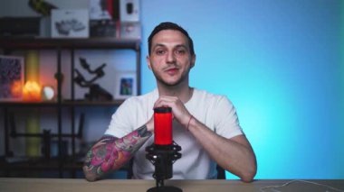 Dövmeli bir Avrupalı erkek sunucu, podcasting veya vloglama için tasarlanmış bir stüdyoda kamera ile görüşlerini paylaşıyor. Temiz kurulum ve kendinden emin teslimat