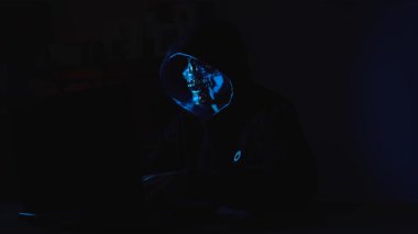  Karanlıkta dizüstü bilgisayar kullanan kapüşonlu bir figür. İsimsiz siber tehditler, online dolandırıcılık ve yeraltı dijital aktiviteleri hakkında güçlü bir kavram..