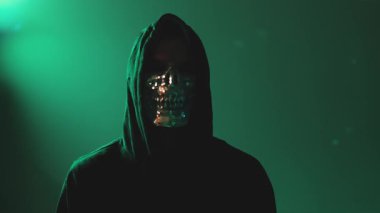 Kapüşonlu maskeli hacker, düşük ışık altında kameraya bakıyor, siber suç ve internet tehdidinin sembolü, gergin ve karanlık ortam