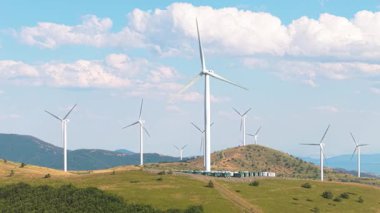 Çimenli yamaçlar ve orman tepeleri üzerinde dönen rüzgar türbinlerinin insansız hava aracı perspektifi. Açık hava çevresel teknoloji başarısını vurguluyor.